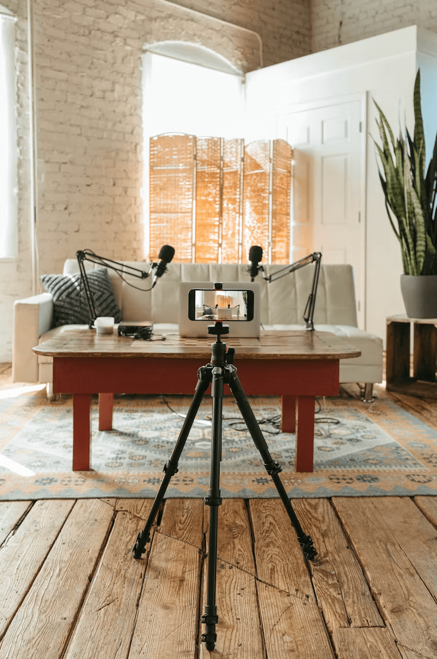 Smartphone on a tripod filming a home podcast studio with microphones and a white sofa.