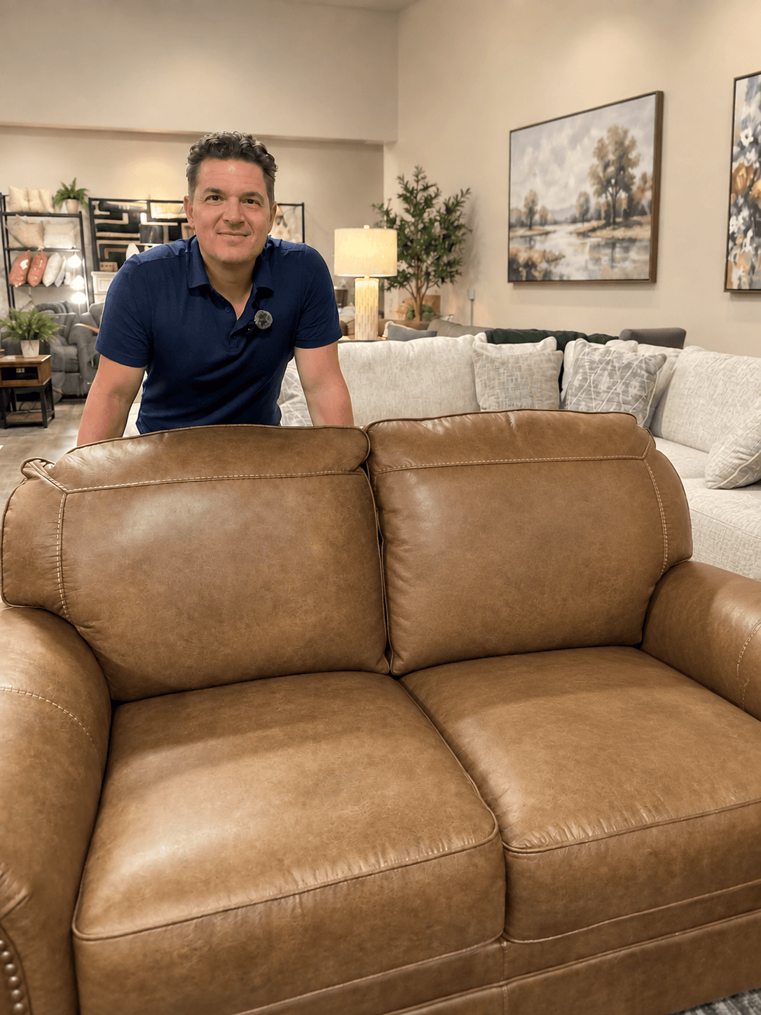 Man in blue polo shirt standing behind a brown leather sofa in a furniture showroom.