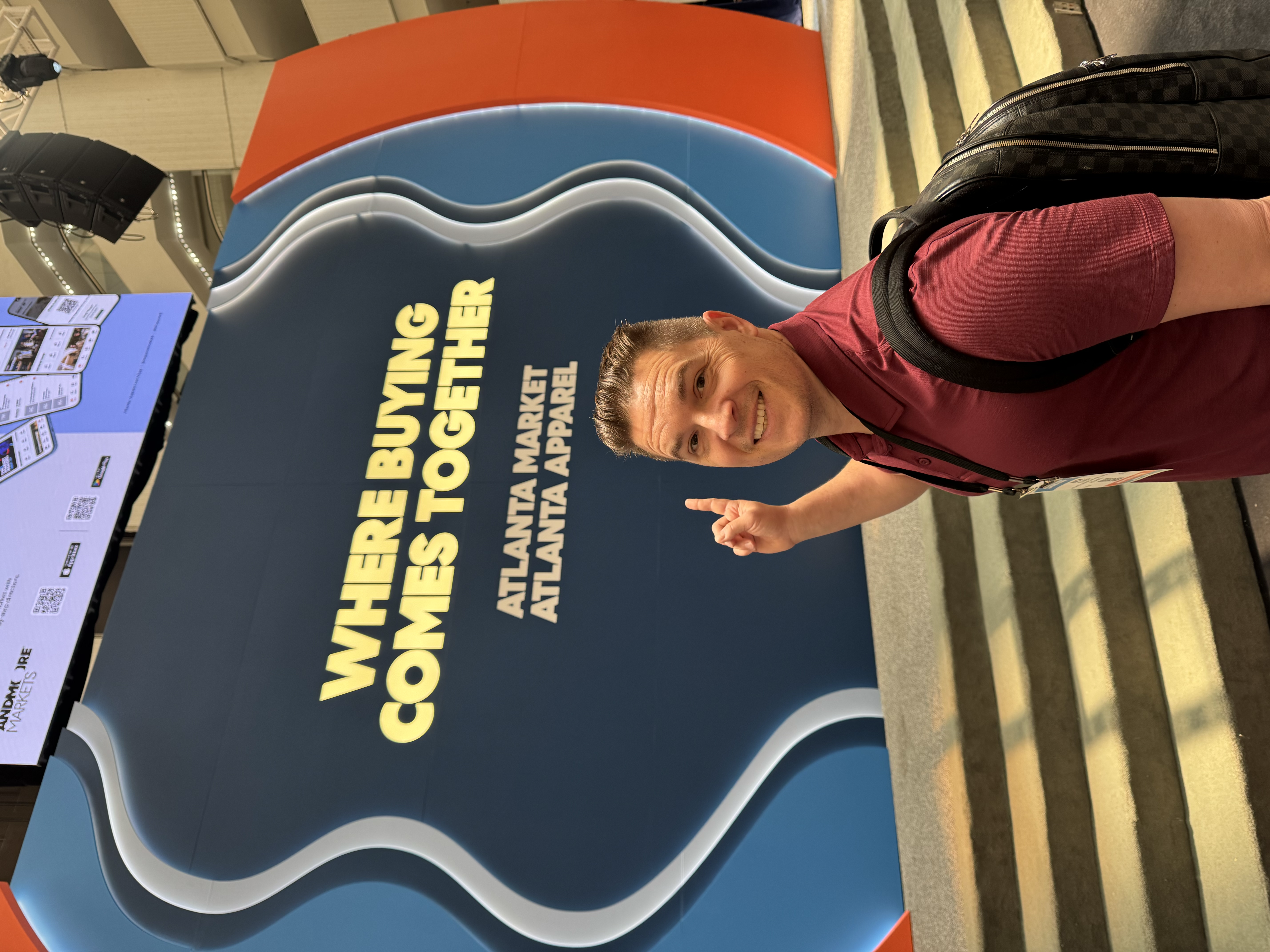 Smiling man in maroon polo points to a sign reading "Where Buying Comes Together".