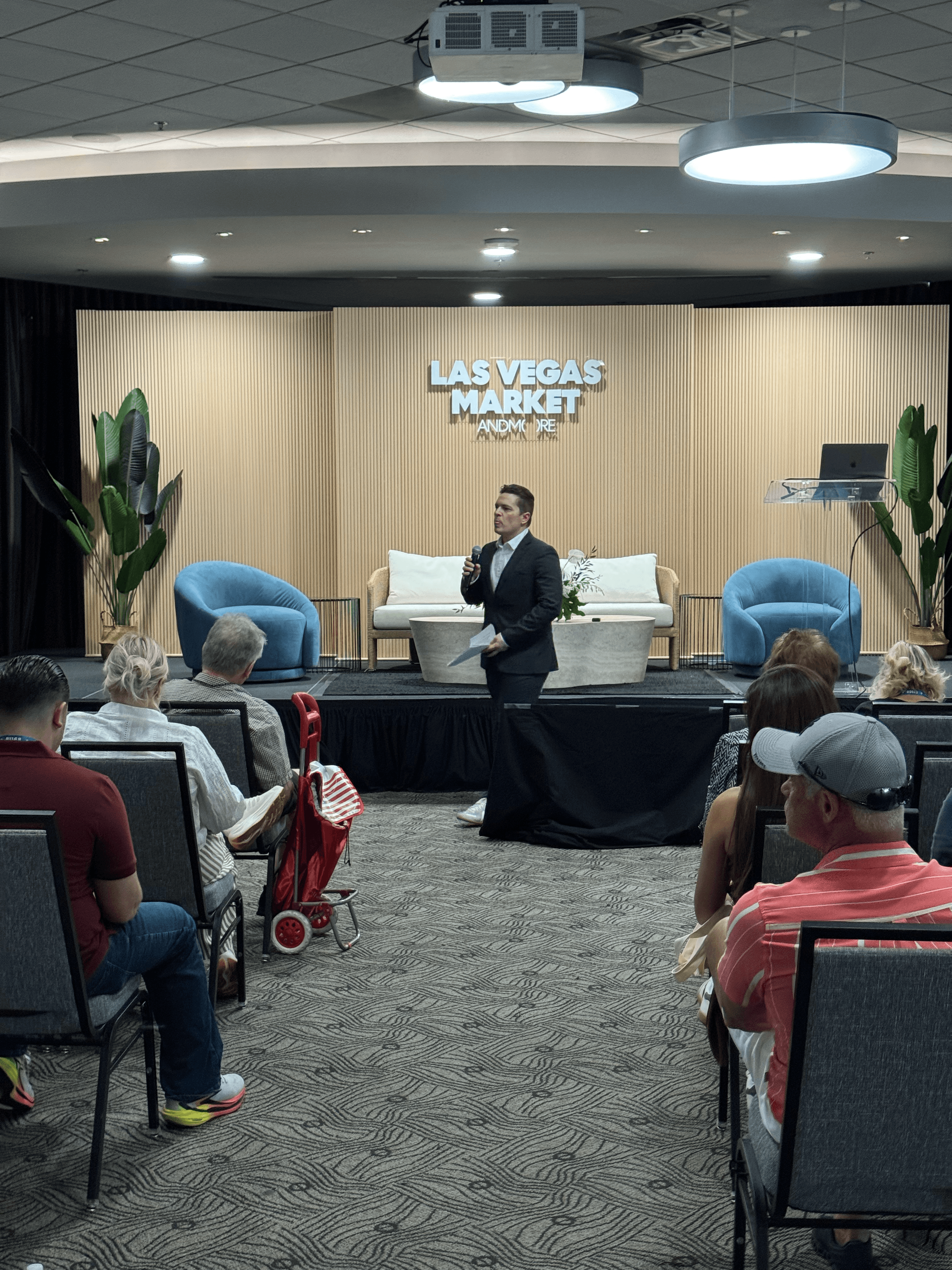 Speaker in a suit presents on stage at Las Vegas Market to a seated audience.